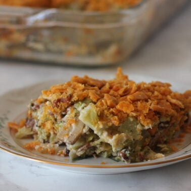 A serving of broccoli casserole, a perfect Thanksgiving side, graces a decorative plate with its crunchy topping. In the background, a glass baking dish brimming with more casserole rests on the marble surface.