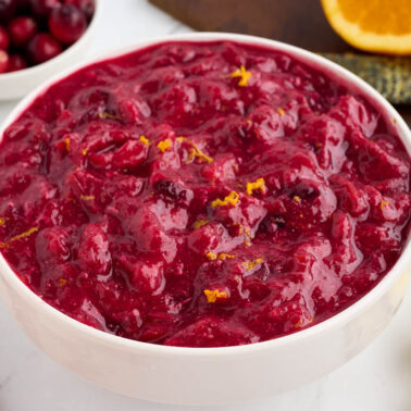 A white bowl filled with cranberry sauce, garnished with orange zest, sits amid cranberries, a slice of orange, and a pineapple wedge—a perfect addition to your Thanksgiving sides.