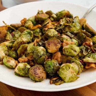 A white plate filled with roasted Brussels sprouts and crispy bacon, making the perfect Thanksgiving side dish. A serving spoon rests inside the dish.