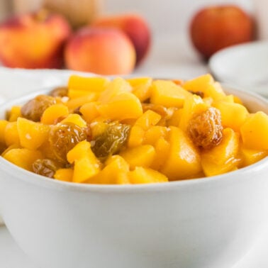 A bowl of diced peaches and raisins sits on a white surface, perfect as a refreshing side dish for Thanksgiving, with whole peaches and plates elegantly arranged in the background.