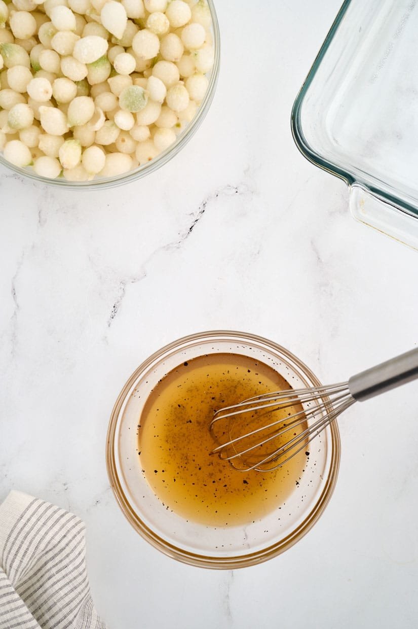 A bowl of dressing with a whisk on a marble surface, next to a bowl of pearl onions and a glass baking dish.