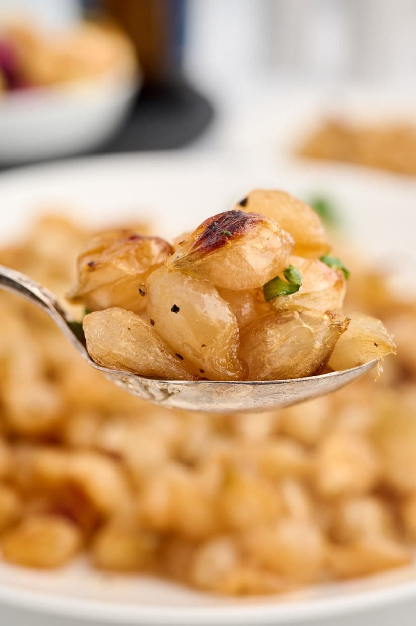 A spoonful of roasted pearl onions, seasoned and garnished, with a plate of onions in the background.