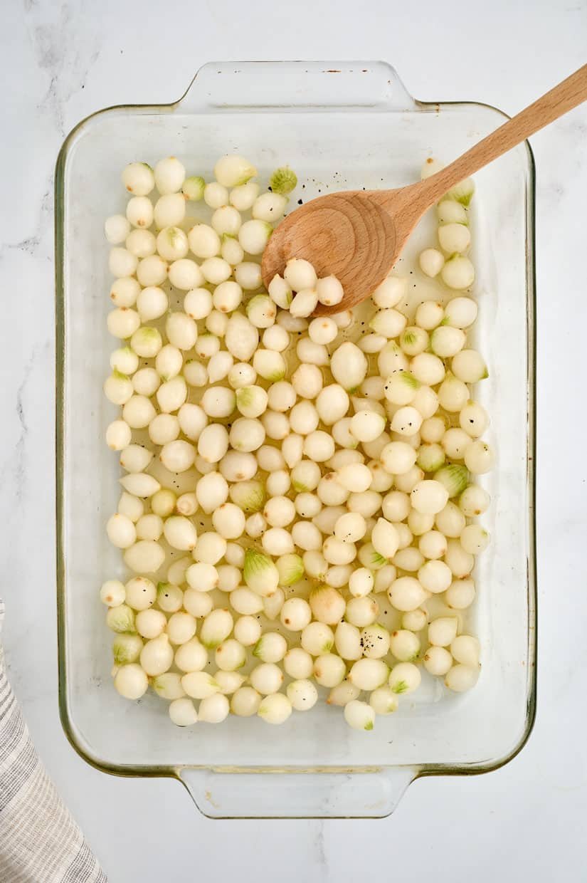 Glass dish with peeled pearl onions, a wooden spoon, and some seasoning.