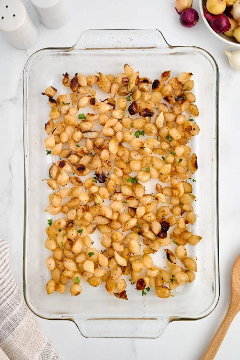 A glass baking dish filled with roasted pearl onions, garnished with herbs, is placed on a white countertop next to a wooden spoon and striped cloth.