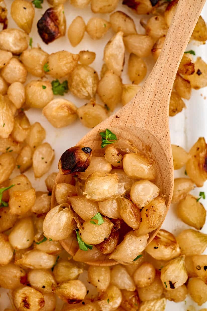 A wooden spoon scoops caramelized roasted pearl onions garnished with parsley.