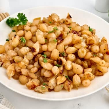 A white plate filled with seasoned and roasted pearl onions, garnished with fresh parsley, set on a light-colored table.