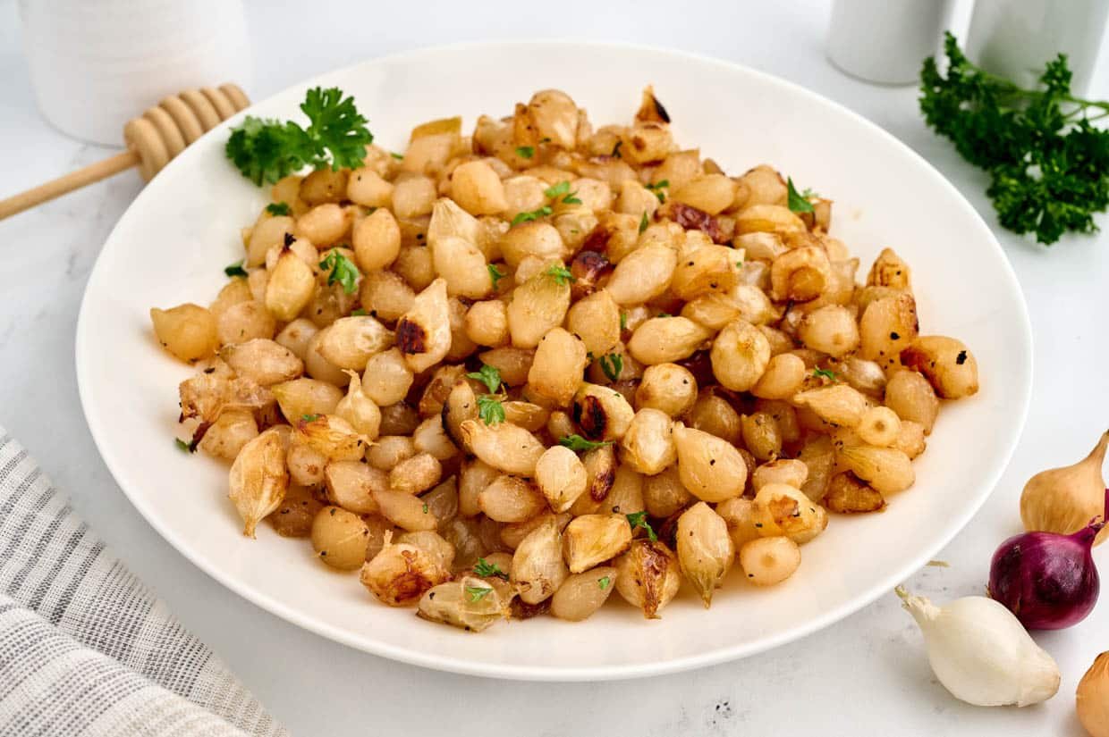 A white plate filled with seasoned and roasted pearl onions, garnished with fresh parsley, set on a light-colored table.