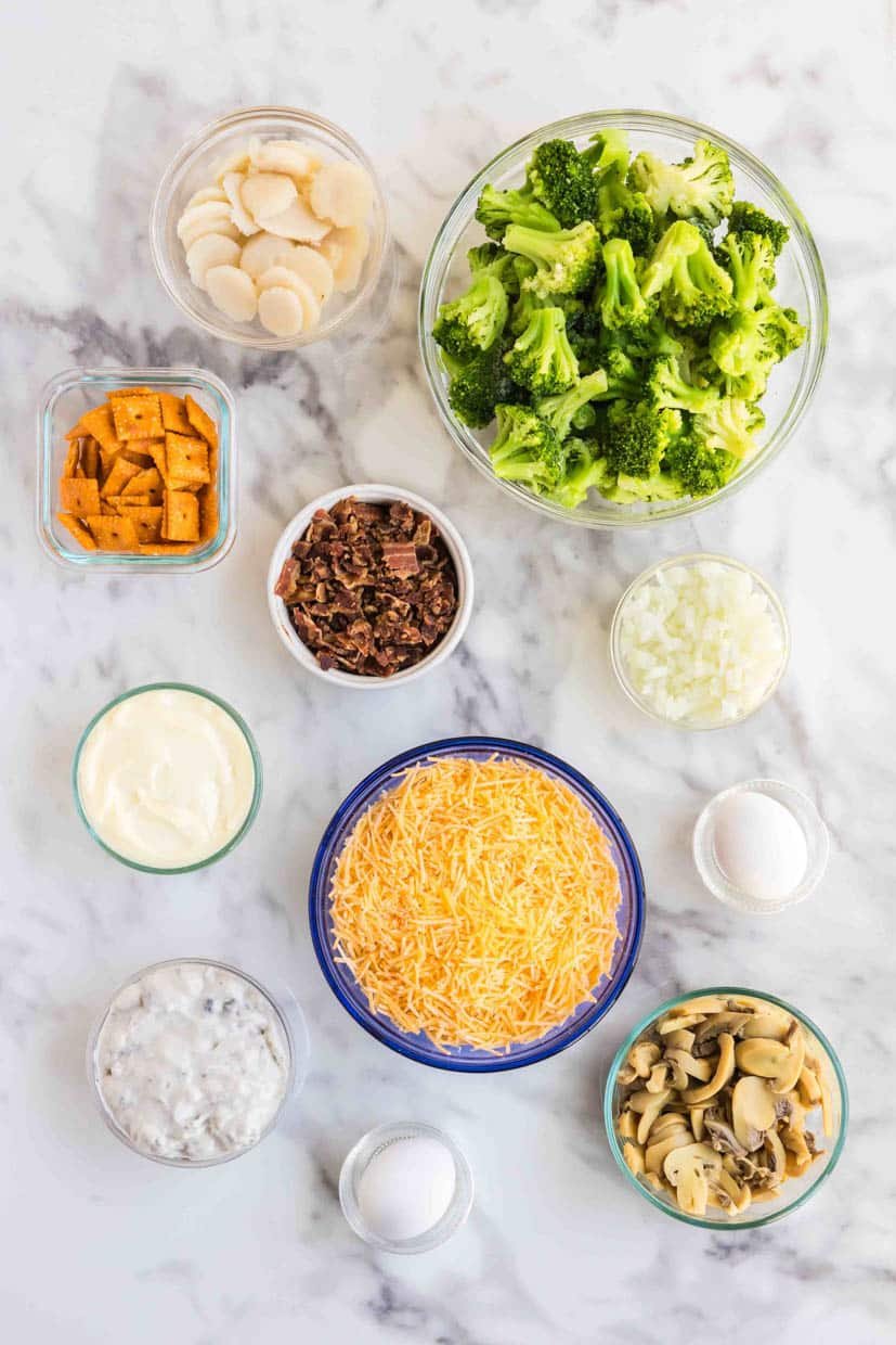 Various bowls containing broccoli, sliced vegetables, grated cheese, diced meat, mayonnaise, and creamy sauces arranged on a marble surface.