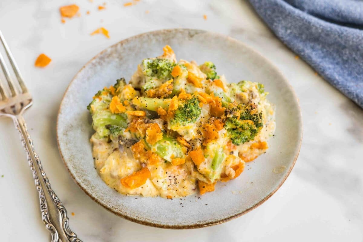 A plate of broccoli cheddar casserole with a creamy sauce, topped with crushed crackers, beside a fork on a marble surface.