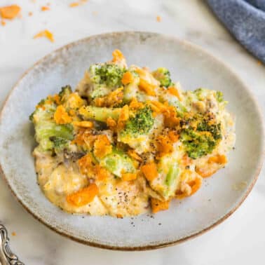A plate of broccoli cheddar casserole with a creamy sauce, topped with crushed crackers, beside a fork on a marble surface.