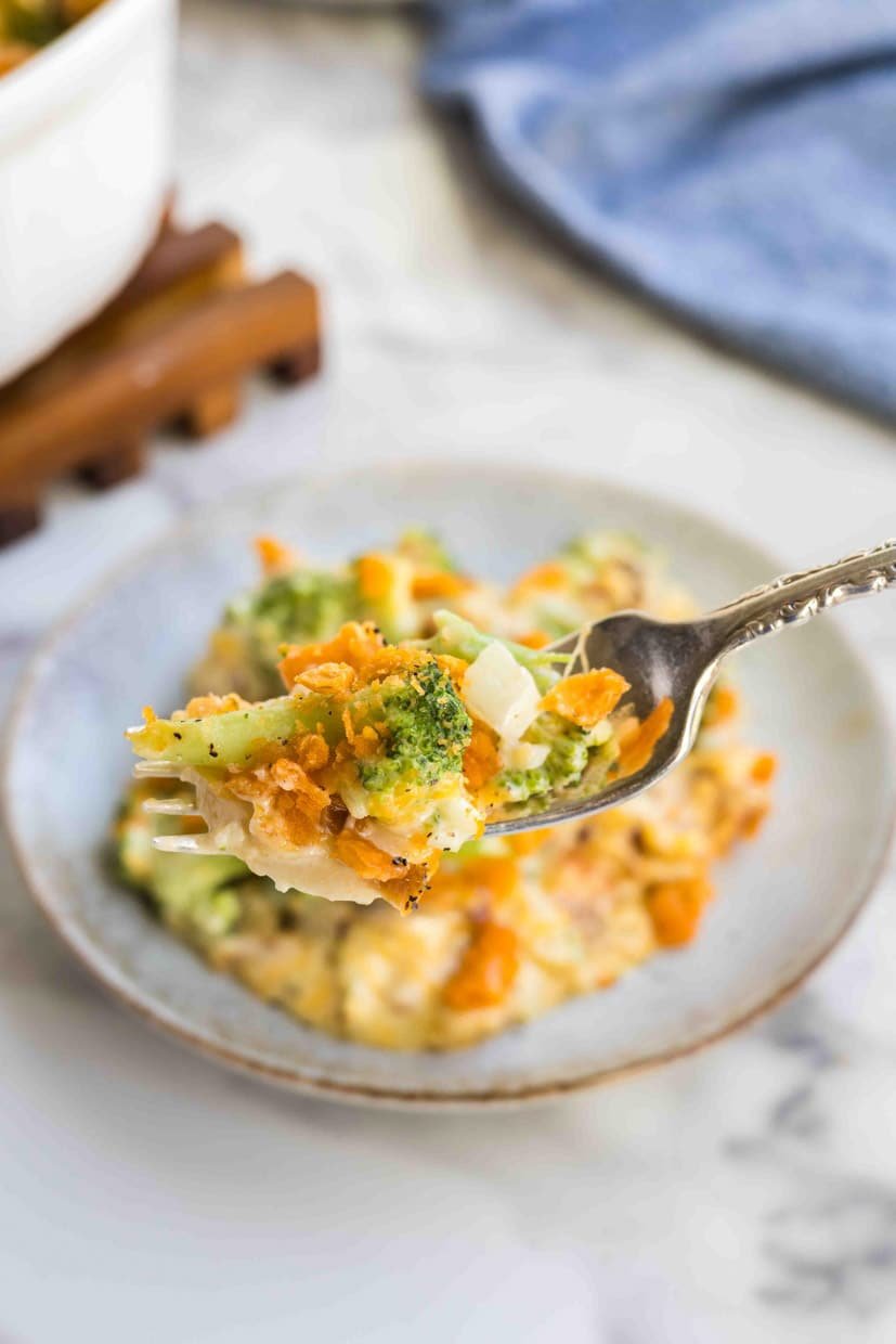 A fork holds a bite of Southern broccoli casserole with visible cheese and breadcrumbs, over a plate with more casserole. A blue cloth is in the background.