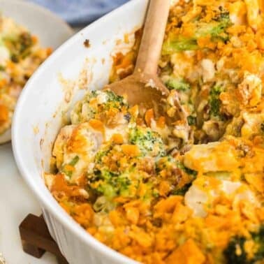 A casserole dish filled with broccoli and cheese, topped with crumbled cheese crackers. A wooden spoon serves a portion onto a plate beside a fork.
