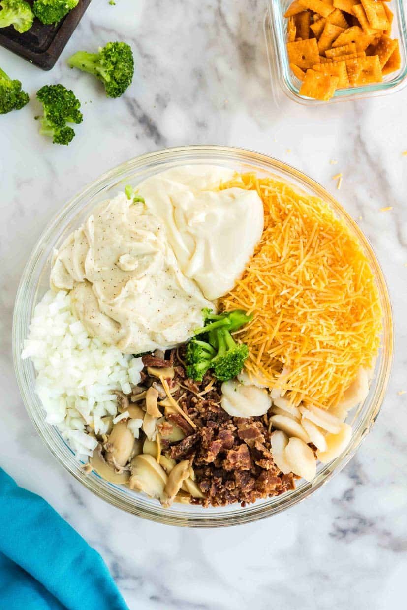 A glass bowl contains chopped onions, broccoli, grated cheese, sliced mushrooms, bacon, and a dollop of dressing on a marble surface, with a blue napkin and snack crackers nearby.