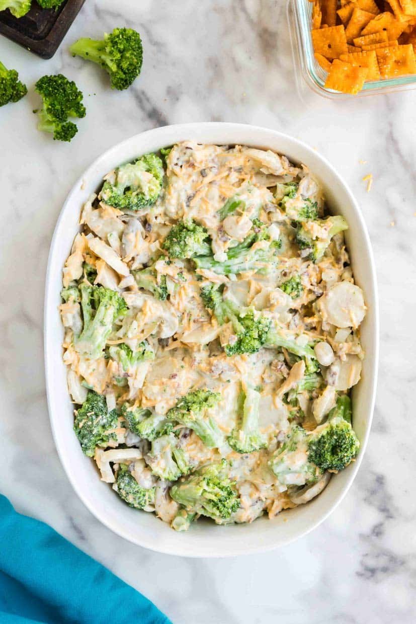 A casserole dish filled with a creamy broccoli and cheese bake on a marble countertop. Nearby are broccoli florets and a container of crackers.