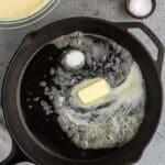 A stick of butter melting in a cast iron skillet with a bowl of batter, a small jug of milk, and a bowl of salt nearby on a gray countertop.