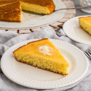 A slice of cornbread with butter on a white plate, with more cornbread on a platter and another slice on a plate in the background.