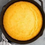 A round skillet with freshly baked golden Southern cornbread on a gray surface, accompanied by a folded white cloth.
