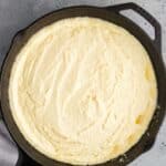 A cast iron skillet filled with a smooth, creamy mixture sits on a gray surface with a cloth partially visible at the bottom.