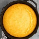 A round skillet containing freshly baked cornbread on a gray surface, with a cloth attached to the handle.