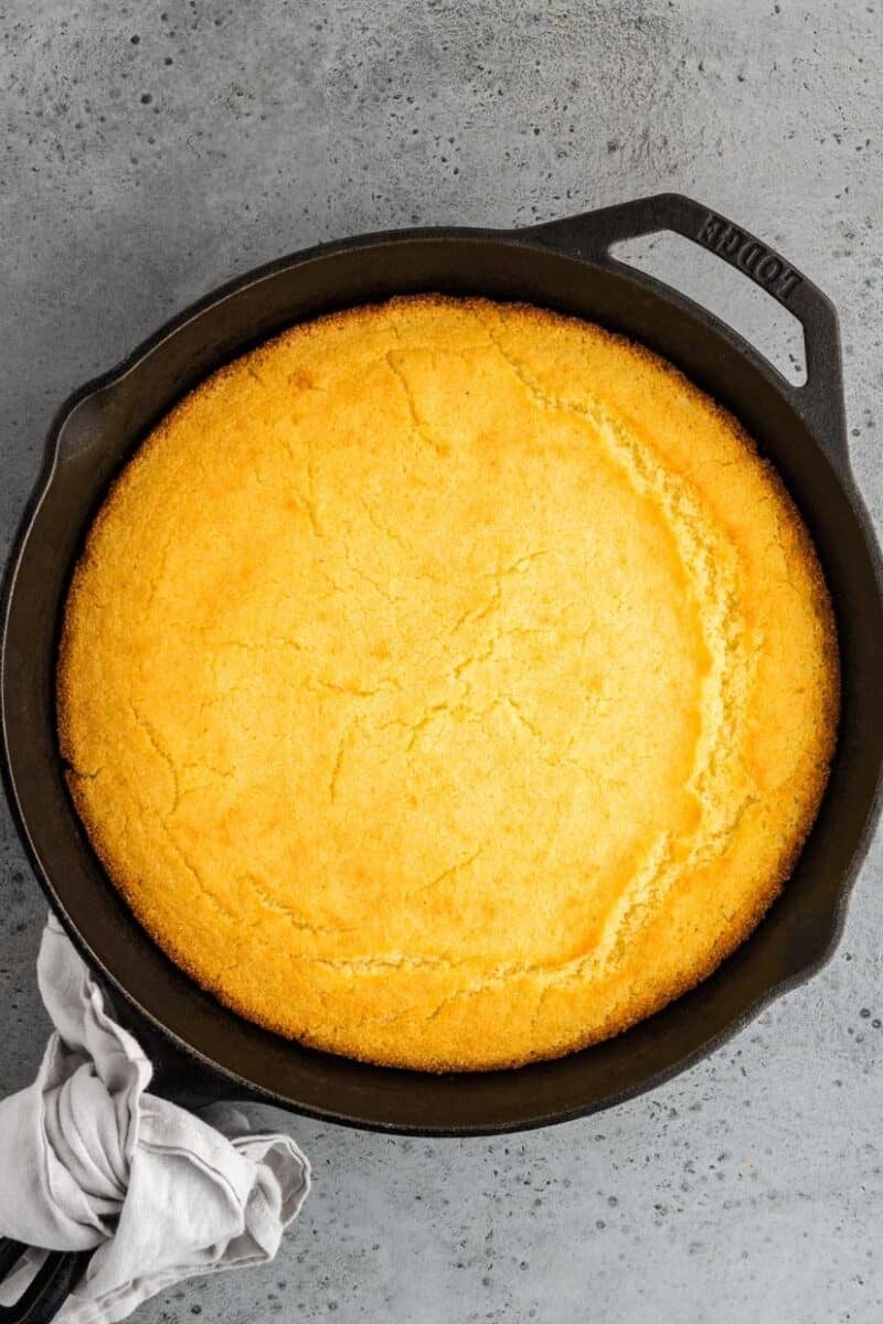A round skillet containing freshly baked cornbread on a gray surface, with a cloth attached to the handle.