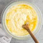 A glass bowl containing a mixed batter with melted butter, being stirred by a wooden spoon. A white cloth lies beside the bowl on a gray surface.