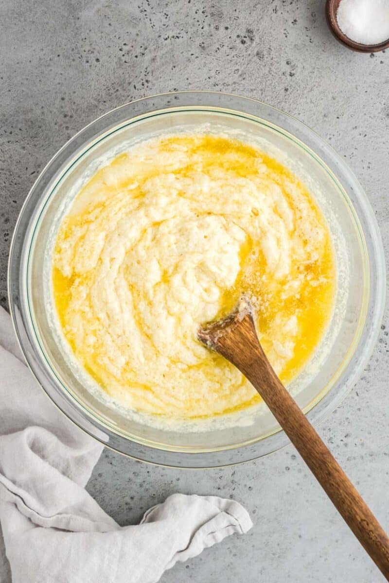 A glass bowl containing a mixed batter with melted butter, being stirred by a wooden spoon. A white cloth lies beside the bowl on a gray surface.