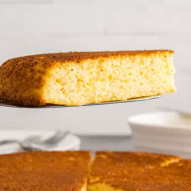 A slice of cornbread is lifted from a pan, with a golden-brown crust and a fluffy yellow interior.