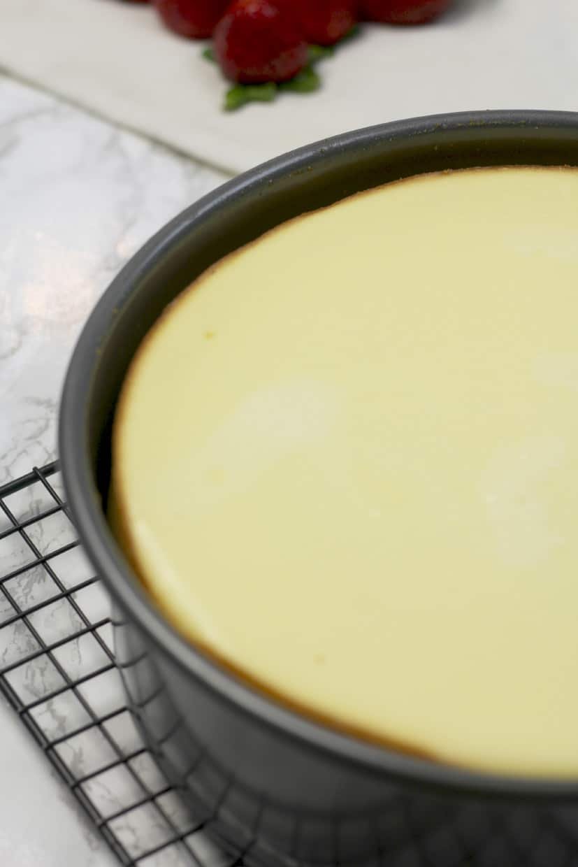 A baked cheesecake in a round pan sits on a cooling rack, with strawberries visible in the background.