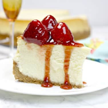 A slice of original cheesecake topped with strawberries and sauce on a white plate. A glass partially visible in the background.