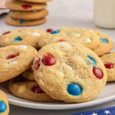 Plate of cookies with red, white, and blue candies, set on a table with an American flag motif, alongside glasses of milk and a stack of cookies in the background.