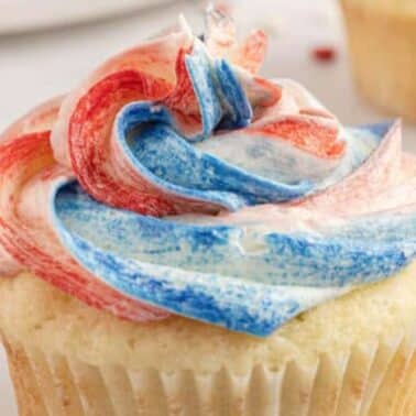 A vanilla cupcake with swirled red, white, and blue frosting. A red patterned napkin and small sprinkles are nearby.