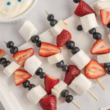 Skewers with strawberries, marshmallows, and blueberries on a white tray next to a bowl of cream topped with blue stars.