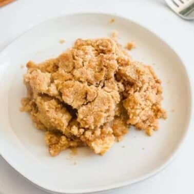 Plate of apple crisp with a fork and cinnamon sticks on a white surface.