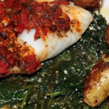 A plate with baked fish topped with tomato sauce, sautéed spinach, and roasted baby potatoes.