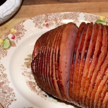 Sliced glazed ham on a decorative plate with a gravy boat beside it on a wooden surface.