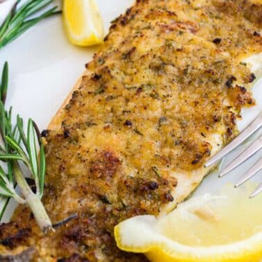 A grilled trout fillet with a golden crust is served on a white plate with rosemary sprigs and lemon wedges, and a fork beside it.