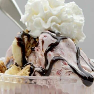 A glass bowl with ice cream topped with whipped cream, chocolate sauce, sliced bananas, and wafer pieces. A spoon is placed in the bowl.