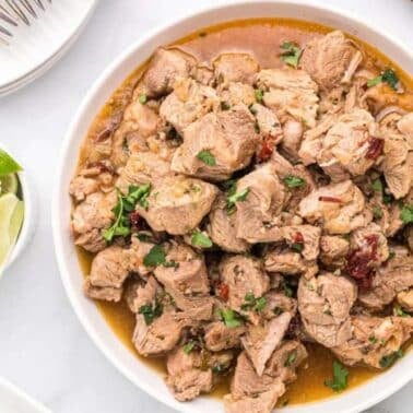 A bowl of cooked braised pork shoulder garnished with herbs, surrounded by lime wedges and tortillas on a table.