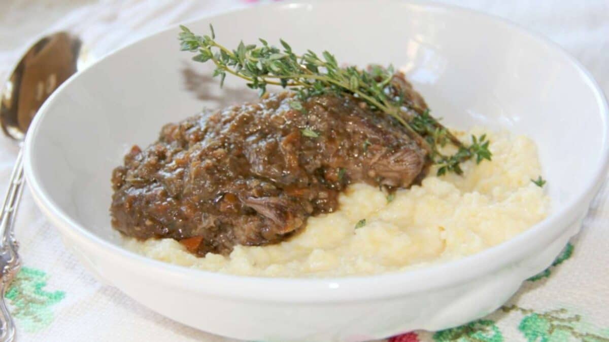 A white bowl of mashed potatoes topped with braised short ribs and gravy, garnished with a sprig of thyme.