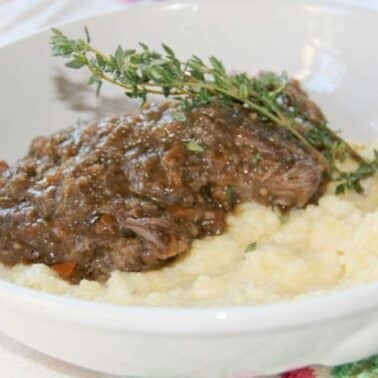 A white bowl of mashed potatoes topped with braised short ribs and gravy, garnished with a sprig of thyme.