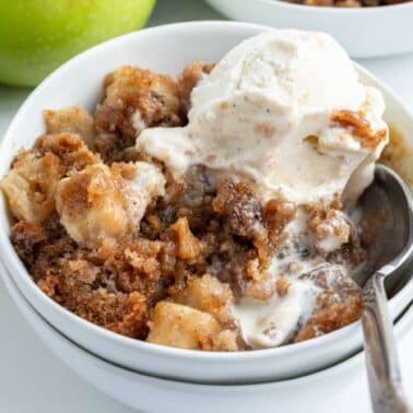 A bowl of caramel apple cake topped with a scoop of vanilla ice cream and a spoon. Green apples and pecans are visible in the background.