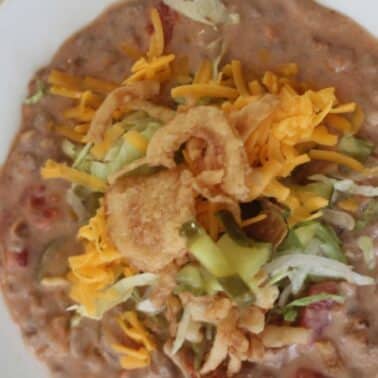 A plate of cheeseburger soup topped with shredded cheddar cheese, lettuce, sliced pickles, and crispy fried onions.