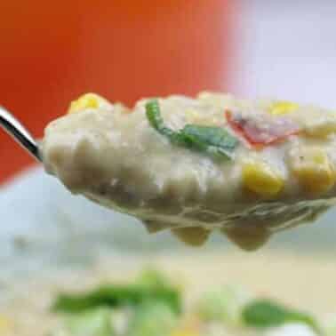A spoonful of creamy chicken corn chowder with corn, green onions, and red peppers held above a bowl.