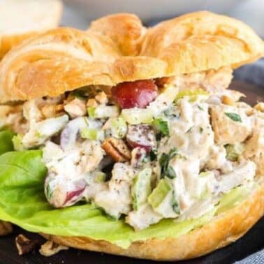 Chicken salad sandwich with lettuce, grapes, chopped nuts, and creamy dressing on a croissant, placed on a dark wooden surface.