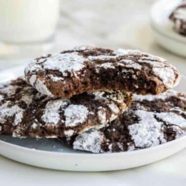 Three chocolate crinkle cookies dusted with powdered sugar on a white plate, with one cookie partially eaten; a glass of milk in the background.