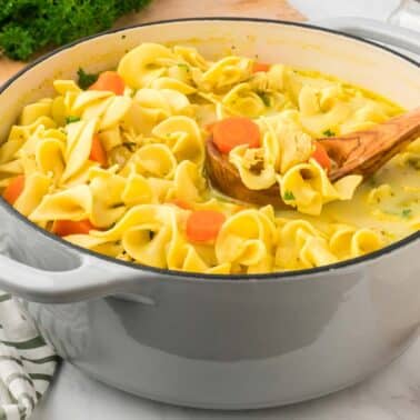 A pot of chicken noodle soup with carrots and parsley, featuring a wooden spoon, on a white countertop with a striped cloth and parsley nearby.