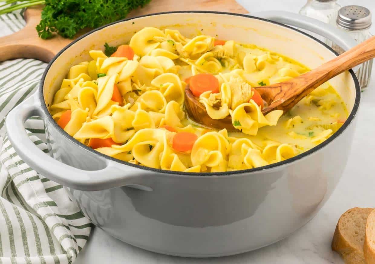 A pot of chicken noodle soup with carrots and parsley, featuring a wooden spoon, on a white countertop with a striped cloth and parsley nearby.