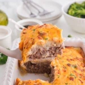 A slice of homemade shepherd's pie with melted cheese is lifted from a white baking dish. Bowls of broccoli and condiments are in the background.