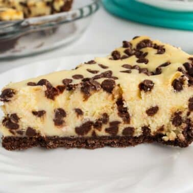A slice of chocolate chip cheesecake on a white plate, featuring a chocolate crust and a creamy filling with chocolate chips. A glass of milk is in the background.