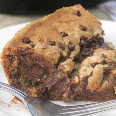 A slice of chocolate chip cookie pie on a white plate with a fork beside it. The pie is thick and filled with chocolate chunks.